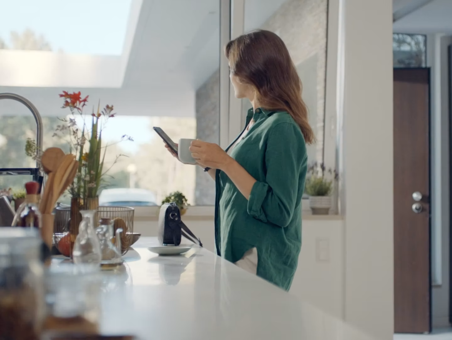 A woman at a kitchen counter, mug in one hand, smartphone in the other, a Hyundai parked in her driveway.